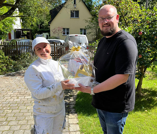 Fabian Sorgalla (rechts) überreicht Maler-Gesellin Jean zur bestandenen Gesellenprüfung draußen bei Sonnenschein einen Geschenkekorb von der Werkstück Raumgestaltung & Versorgungstechnik GmbH in Berlin.