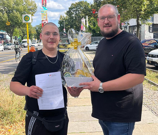 Fabian Sorgalla (rechts) überreicht Geselle Yanneck zur bestandenen Gesellenprüfung draußen bei Sonnenschein einen Geschenkekorb von der Werkstück Raumgestaltung & Versorgungstechnik GmbH in Berlin.
