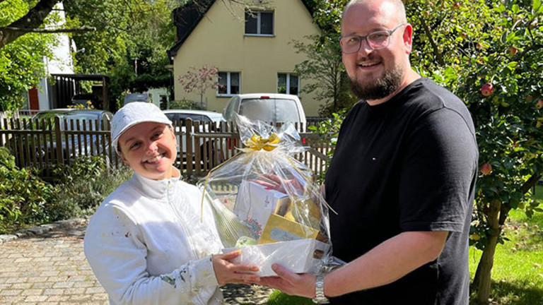Fabian Sorgalla (rechts) überreicht Maler-Gesellin Jean zur bestandenen Gesellenprüfung draußen bei Sonnenschein einen Geschenkekorb von der Werkstück Raumgestaltung & Versorgungstechnik GmbH in Berlin.