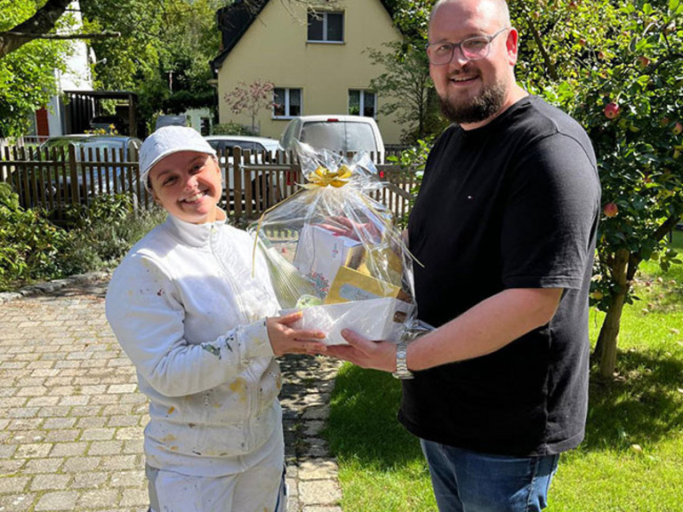 Fabian Sorgalla (rechts) überreicht Maler-Gesellin Jean zur bestandenen Gesellenprüfung draußen bei Sonnenschein einen Geschenkekorb von der Werkstück Raumgestaltung & Versorgungstechnik GmbH in Berlin.