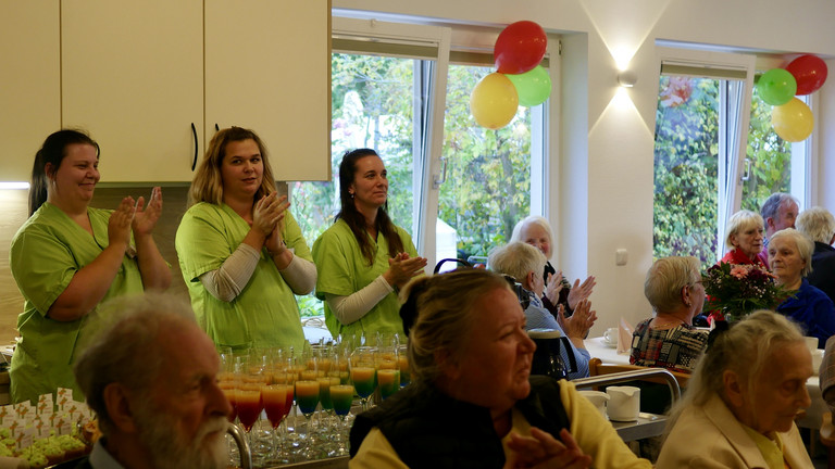 Drei stehende Pflegekräfte in grüner Arbeitskleidung und sitzende Gäste klatschen im bunt geschmückten Saal bei der Wiedereröffnung der Tagespflegestätte für ältere Menschen im Immanuel Seniorenzentrum Kläre Weist in Petershagen Drei stehende Pflegekräfte in grüner Arbeitskleidung und sitzende Gäste klatschen im bunt geschmückten Saal bei der Wiedereröffnung der Tagespflegestätte für ältere Menschen im Immanuel Seniorenzentrum Kläre Weist in Petershagen