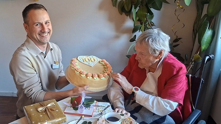 Martin Braungardt überreicht an einem Kaffetisch sitzend eine Torte mit einer verzierten 100 Martin Braungardt überreicht an einem Kaffetisch sitzend eine Torte mit einer verzierten 100