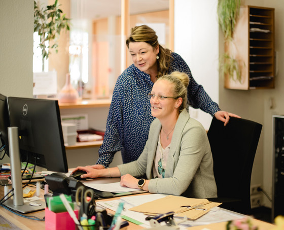 Eine Verwaltungsmitarbeiterin blickt ihrer an einem Schreibtisch sitzenden Kollegin über die Schulter und blickt auf einen Monitor in der Verwaltung der Senioreneinrichtung Immanuel Haus Ecktannen in Waren (Müritz)