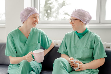 Zwei OP-Fachkräfte mit Kopfhauben und Funktionskleidung sitzen in einem Stationszimmer des Zentral-OP im Albertinen Krankenhaus in Hamburg bei einer Pause mit einer Tasse Kaffee und unterhalten sich 