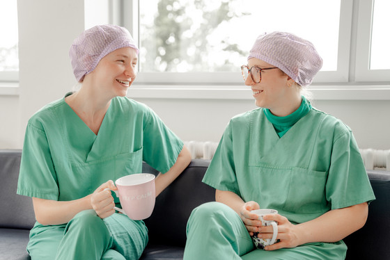 Zwei OP-Fachkräfte mit Kopfhauben und Funktionskleidung sitzen in einem Stationszimmer des Zentral-OP im Albertinen Krankenhaus in Hamburg bei einer Pause mit einer Tasse Kaffee und unterhalten sich 