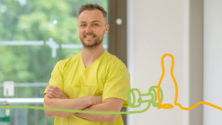 Physiotherapeut steht mit verschränkten Armen in einem Therapiebereich und lächelt in die Kamera. Das Bild steht für professionelle Physiotherapie und Rehabilitation.