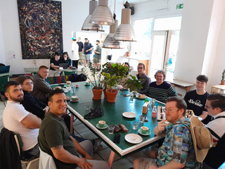 Auszubildende und Ausbildungsverantwortliche sitzen zusammen beim Kaffeetrinken an einem großen Tisch in einem Cafe und blicken Richtung Kamera beim Azubi-Tag 2025 von Werkstück Raumgestaltung und Versorgungstechnik in Berlin