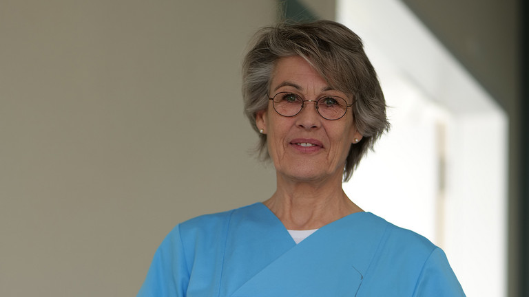Stefanie Heldt, Leitung der Therapeutischen Dienste im Evangelischen Amalie Sieveking Krankenhaus, steht in blauer Arbeitskleidung in einem Klinikflur und blickt in die Kamera.
