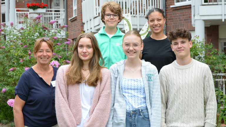 Fünf Schülerinnen und Schülern stehen während ihres Sozialpraktikums mit Pflegedienstleitung Karina Arlt  (links) im Sinnesgarten der Residenz am Wiesenkamp in Hamburg-Volksdorf und lächeln beim Gruppenfoto in die Kamera