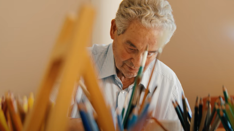 Ein Ehrenamtlicher der Kunsttherapie im Film Gesehen-Fühlen-Leben von Filmemacher Andrew Agbottah über die Menschen im Diakonie Hospiz Wannsee in Berlin sitzt an einem Tisch mit Kunst- und Malutensilien und blickt auf ein Bild