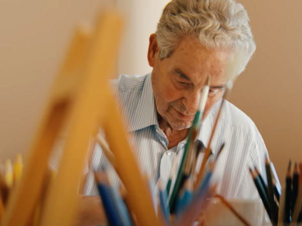 Ein Ehrenamtlicher der Kunsttherapie im Film Gesehen-Fühlen-Leben von Filmemacher Andrew Agbottah über die Menschen im Diakonie Hospiz Wannsee in Berlin sitzt an einem Tisch mit Kunst- und Malutensilien und blickt auf ein Bild