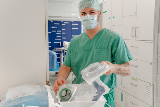 Eine OP-Fachkraft in grüner Funktionskleidung steht in einem Vorbereitungsraum über steril verpackten Materialien für eine Operation und blickt in die Kamera im Zentral-OP im Albertinen Krankenhaus in Hamburg-Schnelsen