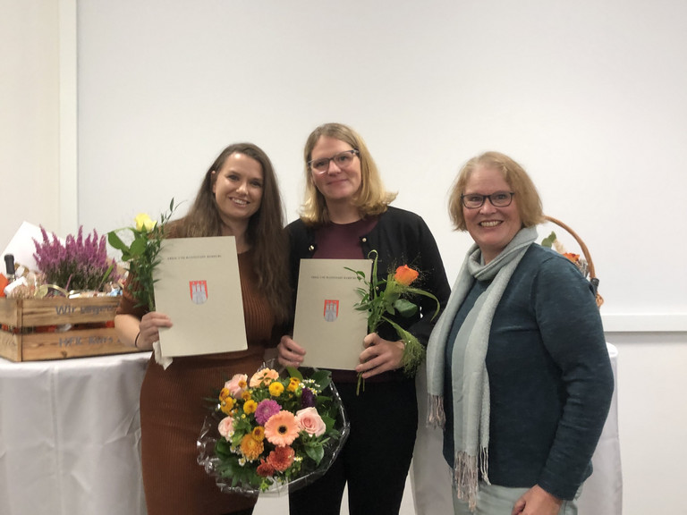 Drei Frauen stehen bei einer Abschlussfeier zusammen; zwei von ihnen halten Zertifikate und Blumensträuße nach bestandener Weiterbildung zur Hygienefachkraft.