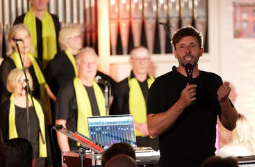 Chorleiter Sören Schröder des Albertinen Mitarbeitenden Gospelchors spricht mit einem Mikrofon in der Hand zum Publikum im festlich beleuchteten Kirchenraum der Kirche am Albertinen Krankenhaus in Hamburg.