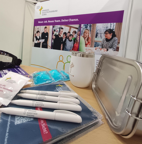 Mit dem Logo der Immanuel Albertinen Diakonie bedruckte Kugelschreiber, eine Brotdose, ein Thermobecher und Infomaterialien auf einem Tisch vor einem großen mit Fotos und Text bedruckten Banner am Messestand des Immanuel Suchthilfeverbunds Guben auf der Ausbildungsmesse Impuls in Cottbus