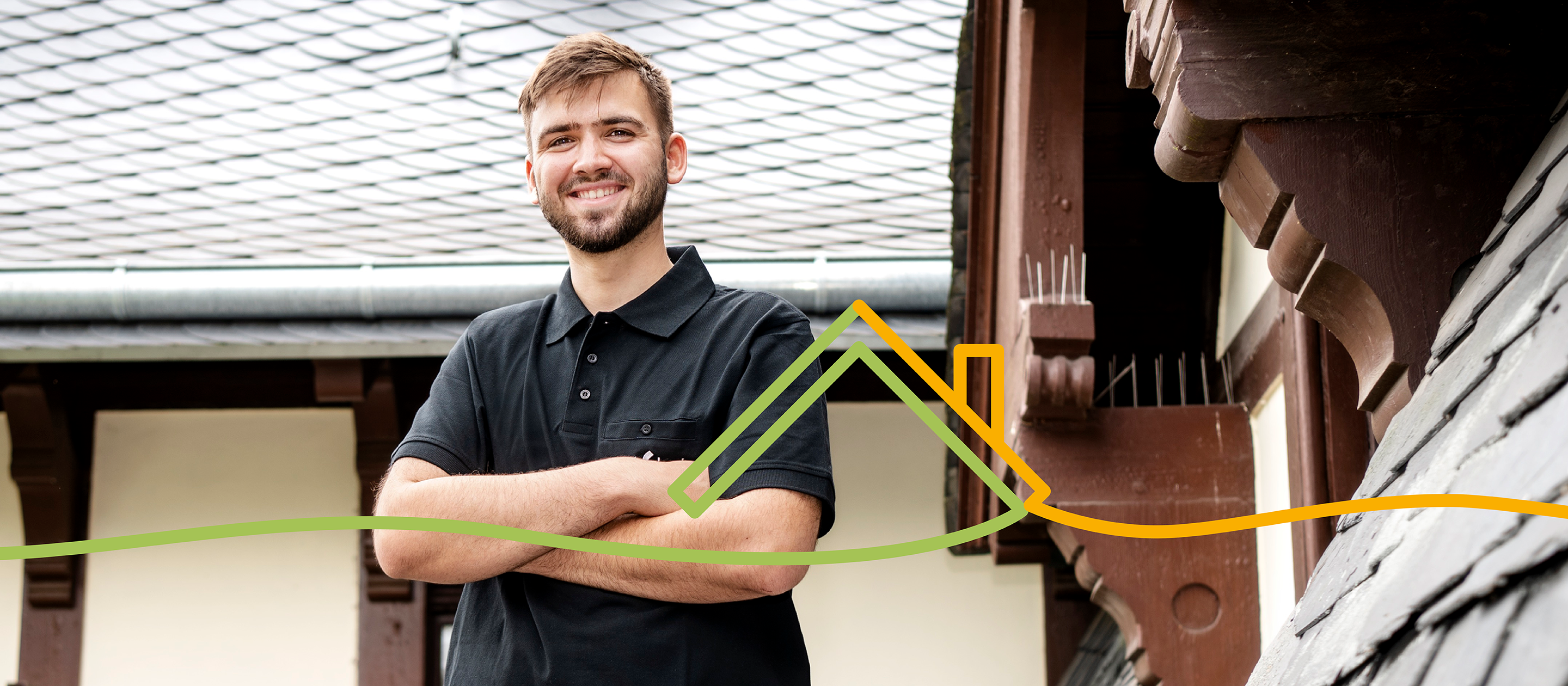 Handwerker steht lächelnd vor traditionellem Gebäude mit Schieferdach – Stellenanzeige im Bereich Dachbau / Werkstück – Arbeiten im Handwerk