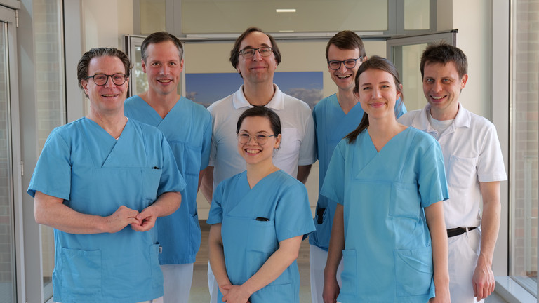 Das Team Des Mitralklappen-Zentrums des Albertinen Herz- und Gefäßzentrums am Standort Hamburg Schnelsen lächelt auf einem Flur in Arztkleidung in die Kamera