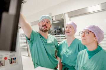 Drei OP-Fachkräfte mit Kopfhauben und grüner Funktionskleidung stehen in einem Stationsflur und blicken auf einen Monitor an der Wand im Zentral-OP des Albertinen Krankenhauses in Hamburg-Schnelsen