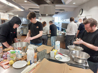 Drei Auszubildende stehen um einen Tisch mit Küchenutensilien wie Schüsseln, Mehl und weiteren Zutaten und kneten Plätzchenteig in der Küche der Gemeinde der Baptisten in Berlin-Schönberg beim 4. Azubitag der Auszubildenden von Werkstück Berlin mit Unterstützung von Immanuel Dienstleistungen