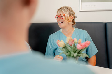 Eine Krankenpflegerin in grünblauer Funktionskleidung sitzt in einer Sitzecke mit Blumen auf dem Tisch und unterhält sich fröhlich mit Personen im Hintergrund im Albertinen Krankenhaus in Hamburg-Schnelsen