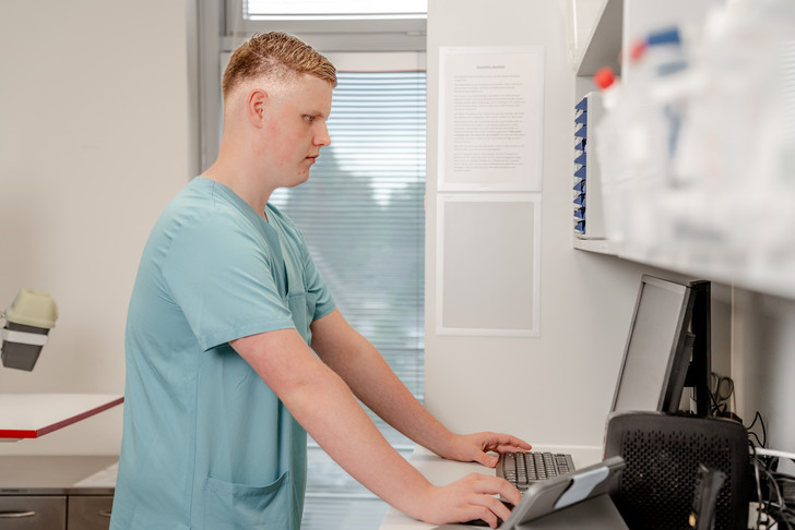 Ein jüngerer Pfleger in grünblauer Funktionskleidung steht in einem Stationszimmer und arbeitet an einem Computer im Albertinen Krankenhaus in Hamburg-Schnelsen Ein jüngerer Pfleger in grünblauer Funktionskleidung steht in einem Stationszimmer und arbeitet an einem Computer im Albertinen Krankenhaus in Hamburg-Schnelsen