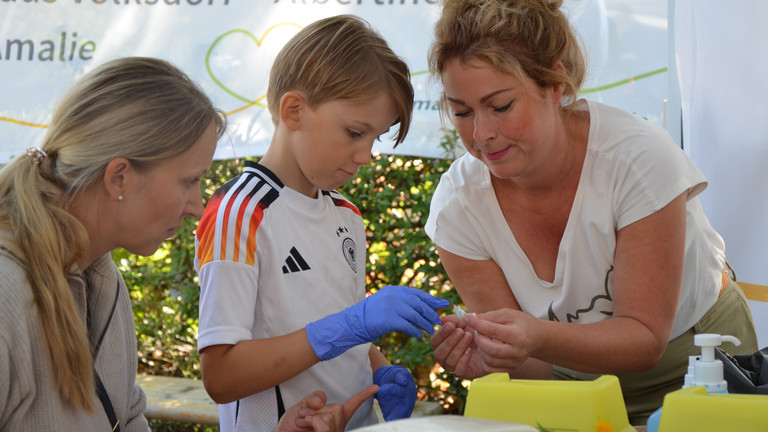 Ein Kind mit blauen Einmalhandschuhen bei einer Simulationsübung mit Mitarbeiterinnen an den Ständen des Gesundheitscampus Volksdorf der Immanuel Albertinen Diakonie beim Stadtteilfest Volksdorf in Hamburg