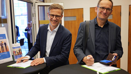 Zwei Gäste tragen sich am Empfang in eine Liste ein und blicken in die Kamera beim Einführungssymposium „Die Zukunft ist alt!“ von Priv.-Doz. Dr. Gödel, Chefarzt der Klinik für Geriatrie im Albertinen Zentrum für Altersmedizin im Albertinen Krankenhaus in Hamburg.