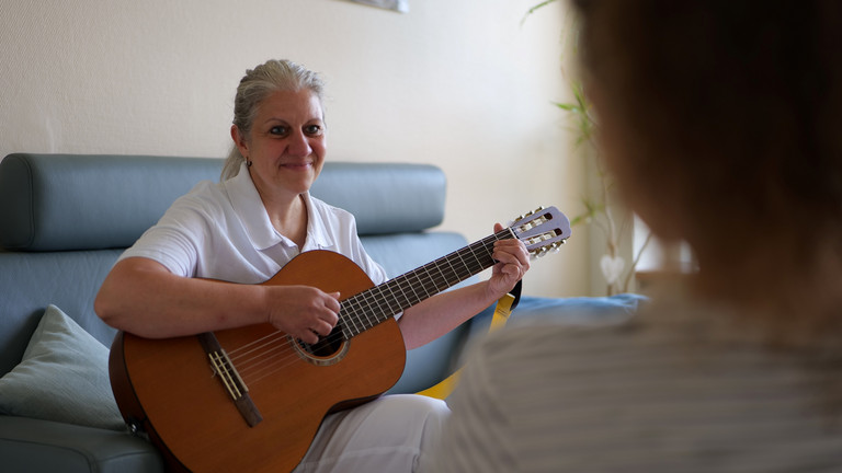 Eine Musiktherapeutin der Palliativeinheit sitzt mit einer Gitarre auf einem Sofa und blickt in Richtung einer Patientin im Interdisziplinären Therapiezentrum im Albertinen Krankenhaus Hamburg Eine Musiktherapeutin der Palliativeinheit sitzt mit einer Gitarre auf einem Sofa und blickt in Richtung einer Patientin im Interdisziplinären Therapiezentrum im Albertinen Krankenhaus Hamburg