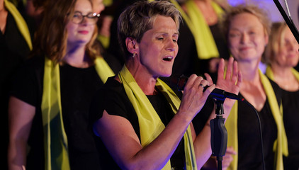 Eine Sängerin steht mit einem grünen Schal beim Solo am Mikrofon auf der Bühne neben anderen Sängerinnen und Sängern des Albertinen Mitarbeitenden Gospelchors beim Konzert in der Kirche am Albertinen Krankenhaus in Hamburg. 
