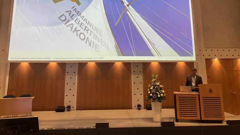 Der Leiter der Perfusiologie am Immanuel Klinikum Bernau Herzzentrum Brandenburg Torsten Müller stellt den Perfusionscontainer am Rednerpult auf der Bühne mit einer Leinwand bei der Internationalen Jahrestagung der Deutschen Gesellschaft für Perfusionsmedizin und der Fokustagung Herz der Deutschen Gesellschaft für Thorax-, Herz- und Gefäßchirurgie in Leipzig vor