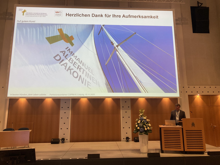 Der Leiter der Perfusiologie am Immanuel Klinikum Bernau Herzzentrum Brandenburg Torsten Müller stellt den Perfusionscontainer am Rednerpult auf der Bühne mit einer Leinwand bei der Internationalen Jahrestagung der Deutschen Gesellschaft für Perfusionsmedizin und der Fokustagung Herz der Deutschen Gesellschaft für Thorax-, Herz- und Gefäßchirurgie in Leipzig vor