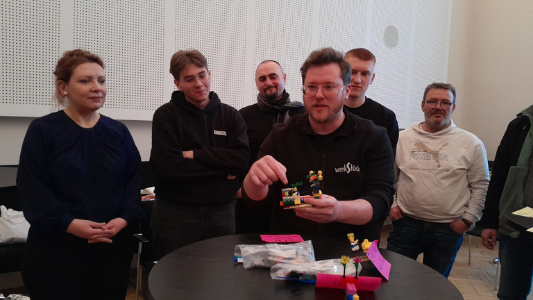 Ein Mann spricht zur umstehenden Gruppe an einem Tisch mit einem Segelboot aus bunten Legosteinen beim Teamtag vom Meisterbetrieb für Handwerk Werkstück Berlin zum Thema "Gute Kommunikation verbindet" in Berlin-Wannsee Ein Mann spricht zur umstehenden Gruppe an einem Tisch mit einem Segelboot aus bunten Legosteinen beim Teamtag vom Meisterbetrieb für Handwerk Werkstück Berlin zum Thema "Gute Kommunikation verbindet" in Berlin-Wannsee