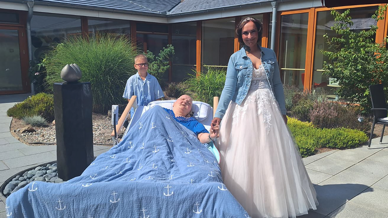 Gästin des Albertinen Hospiz Norderstedt hält im sonnigen Innenhof die Hand ihrer Tochter im Hochzeitskleid; ihr Enkel steht hinter dem Bett.
