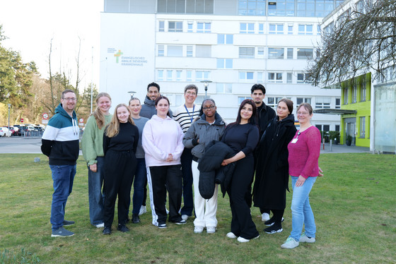 Eine Gruppe neuer Auszubildenden für Gesundheitsberufe steht auf einer grünen Wiese vor dem Evangelischen Amalie Sieveking Krankenhaus und blickt gemeinsam mit zwei hauptamtlichen Praxisanleiterinnen und einem Praxisanleiter in die Kamera.