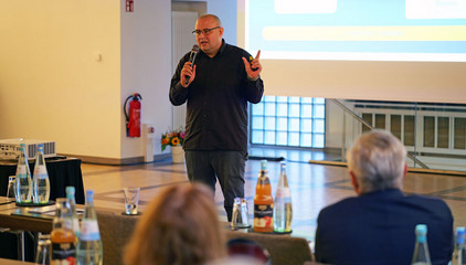 Dr. Schöps spricht mit einem Mikrofon in der Hand neben einer Projektionsleinwand zum Publikum bei einem Vortrag beim Einführungssymposium „Die Zukunft ist alt!“ von Priv.-Doz. Dr. Gödel, Chefarzt der Klinik für Geriatrie im Albertinen Zentrum für Altersmedizin im Albertinen Krankenhaus in Hamburg.