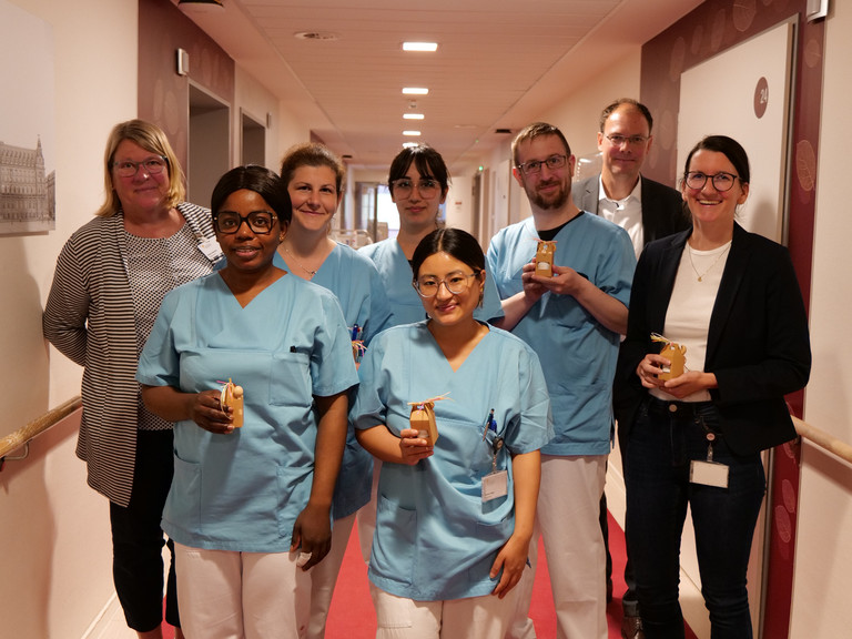 Mehrere Pflegekräfte in blauer Funktionskleidung halten kleine Präsente in den Händen und stehen mit Pflegedirektorin Maike Sellentin, ihrer Stellvertretrin Sabrina Oberle und Krankenhausgeschäftsführer Andreas Schneider in einem Krankenhausflur zum Internationalen Tag der Pflegenden 2026. 