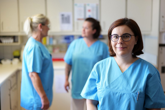 Eine Pflegerin blickt im Stationszimmer der Inneren Medizin des Albertinen Krankenhauses in Hamburg-Schnelsen in die Kamera, im Hintergrund unterhalten sich zwei Pflegekräfte