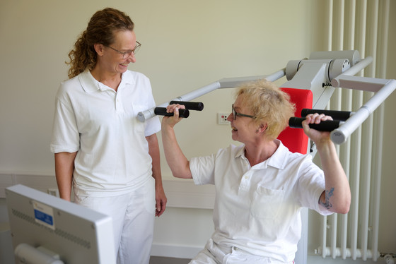 Eine geriatrische Therapeutin sitzt in weißer Funktionskleidung an einem Trainingsgerät für Rücken und Schulter und spricht mit einer danebenstehenden Kollegin im Interdisziplinären Therapiezentrum im Albertinen Krankenhaus Hamburg