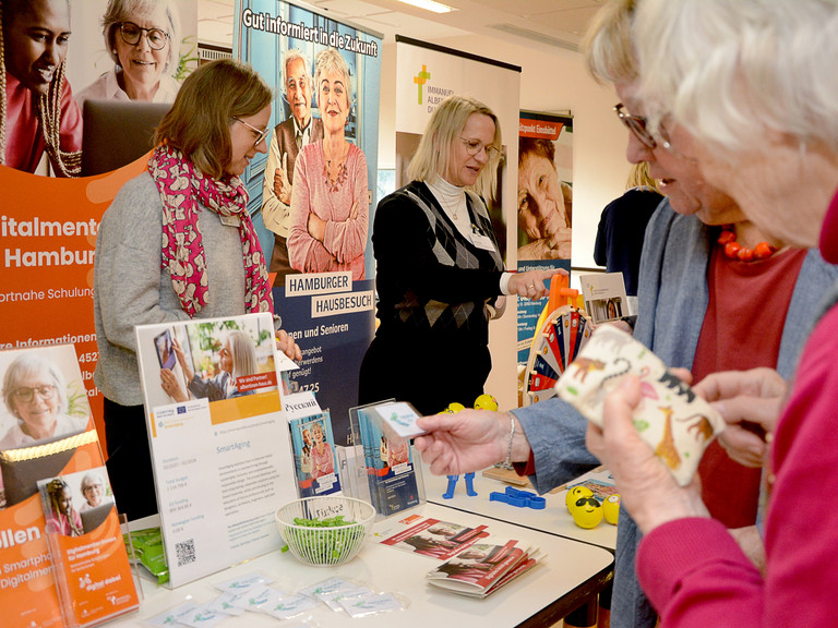 Zwei Mitarbeitende stehen vor Informationsplakaten am Infostand Albertinen Haus auf dem Eimsbütteler Seniorentag, während zwei Frauen die ausgelegten Informationsmaterialien und Giveaways auf dem Tisch näher betrachten Zwei Mitarbeitende stehen vor Informationsplakaten am Infostand Albertinen Haus auf dem Eimsbütteler Seniorentag, während zwei Frauen die ausgelegten Informationsmaterialien und Giveaways auf dem Tisch näher betrachten