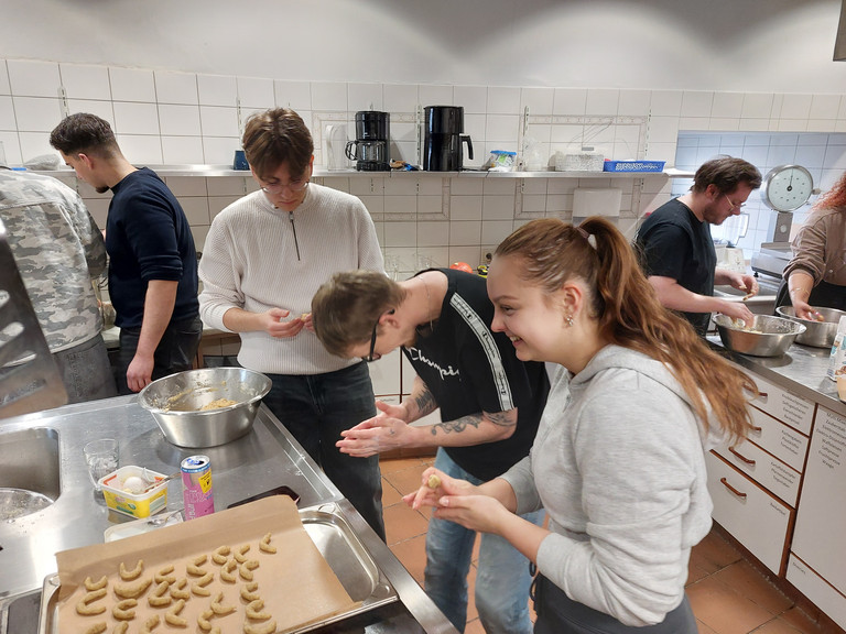 Drei junge Auszubildende stehen um einem Tisch mit einem Backblech und Schüsseln, unterhalten sich und kneten Plätzchenteig in der Küche der Gemeinde der Baptisten in Berlin-Schönberg beim 4. Azubitag der Auszubildenden von Werkstück Berlin mit Unterstützung von Immanuel Dienstleistungen