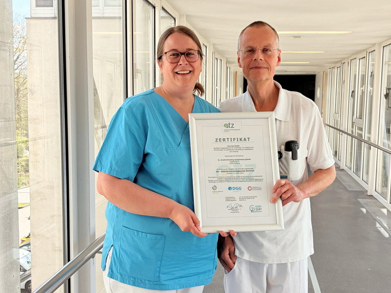 Dr. Saskia Otte, Chefärztin der Geriatrie und Physikalischen Medizin, und Dr. Olaf Pingen, Chefarzt der Orthopädie und Unfallchirurgie, stehen im Amalie Sieveking Krankenhaus in Hamburg-Volksdorf in einem hellen Durchgang und halten gemeinsam das Zertifikat für die erfolgreiche Zertifizierung des Krankenhauses als Alterstraumatologisches Zentrum in die Kamera. 
