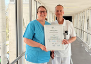 Dr. Saskia Otte, Chefärztin der Geriatrie und Physikalischen Medizin, und Dr. Olaf Pingen, Chefarzt der Orthopädie und Unfallchirurgie, stehen im Amalie Sieveking Krankenhaus in Hamburg-Volksdorf in einem hellen Durchgang und halten gemeinsam das Zertifikat für die erfolgreiche Zertifizierung des Krankenhauses als Alterstraumatologisches Zentrum in die Kamera. 