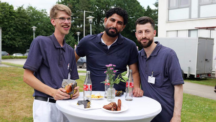 Drei Mitarbeiter stehen in Funktionskleidung an einem Stehtisch mit Blumenschmuck und Getränken und blicken in die Kamera beim Sommerfest 2026 auf dem Gesundheitscampus Volksdorf des Evangelischen Amalie Sieveking Krankenhauses, der Residenz am Wiesenkamp, des Feierabendhauses Volksdorf und der Albertinen Kindertagesstätte Volksdorf. 