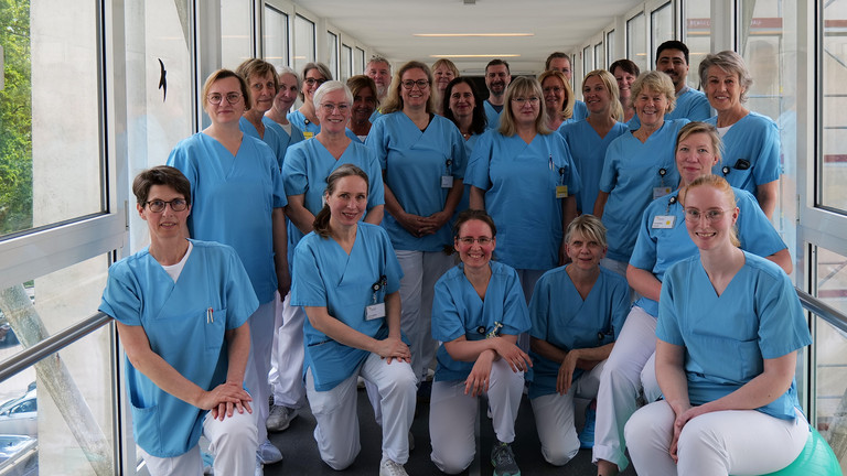 Das Team aus dem Bereich therapeutische Dienste, Geriatrie und Physikalische Medizin im Evangelischen Amalie Sieveking Krankenhaus steht für ein Gruppenfoto zusammen in blauer Funktionskleidung in einen Verbindungsgang im Evangelischen Amalie Sieveking Krankenhaus in Hamburg.