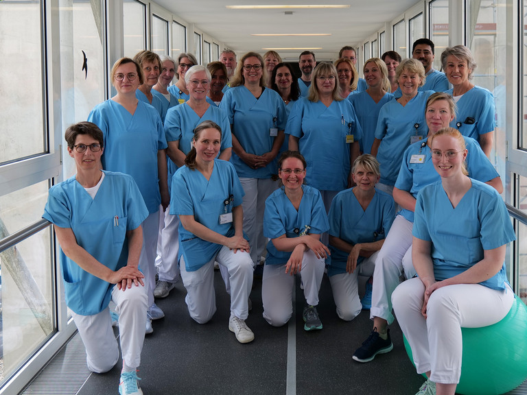 Das Team aus dem Bereich therapeutische Dienste, Geriatrie und Physikalische Medizin im Evangelischen Amalie Sieveking Krankenhaus steht für ein Gruppenfoto zusammen in blauer Funktionskleidung in einen Verbindungsgang im Evangelischen Amalie Sieveking Krankenhaus in Hamburg.