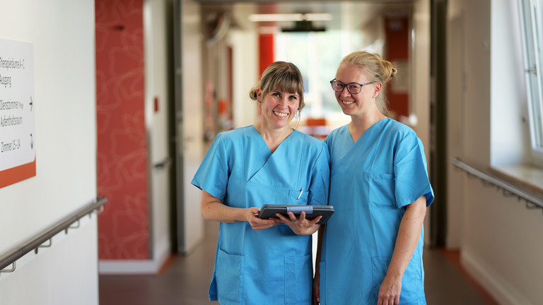 Zwei Pflegefachkräfte in Funktionskleidung stehen mit einem Tablet in den Händen auf einem Stationsflur und blicken in die Kamera im Albertinen Zentrum für Altersmedizin in Hamburg-Schnelsen