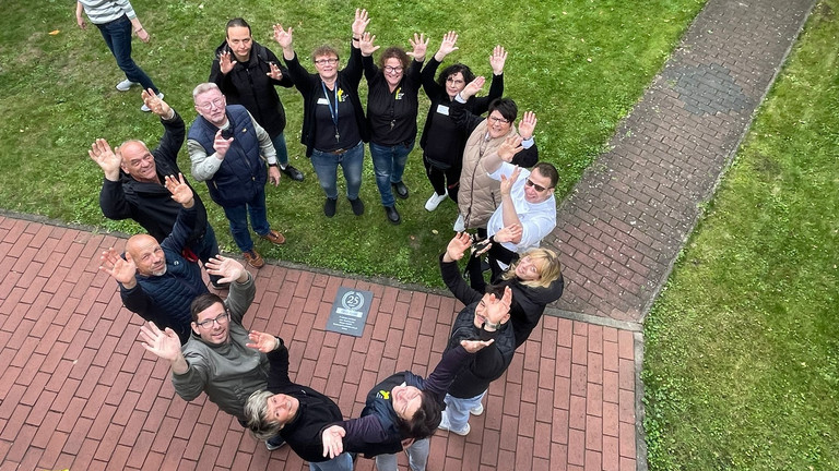 Ein Team von Mitarbeitenden steht im Kreis und blickt nach oben die Kamera zum 25-jährigen Jubiläum des Immanuel Suchthilfeverbunds Guben mit besonderen Wohnformen für abhängigkeitskranke Menschen in Thüringen Ein Team von Mitarbeitenden steht im Kreis und blickt nach oben die Kamera zum 25-jährigen Jubiläum des Immanuel Suchthilfeverbunds Guben mit besonderen Wohnformen für abhängigkeitskranke Menschen in Thüringen