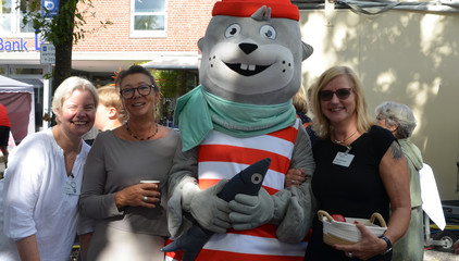 Drei Besucherinnen mit einem Maskottchen blicken in die Kamera bei den Ständen des Gesundheitscampus Volksdorf der Immanuel Albertinen Diakonie beim Stadtteilfest Volksdorf in Hamburg