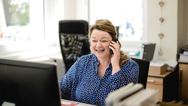 Eine Verwaltungsmitarbeiterin sitzt an einem Schreibtisch und blickt mit einem Telefonhörer in der Hand auf einen Monitor in der Verwaltung der Senioreneinrichtung Immanuel Haus Ecktannen in Waren (Müritz)