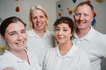 Das Team der Physiotherapie lächelt in weiß gekleidet in die Selfiekamera vor der Kletterwand im Albertinen Krankenhaus Hamburg Schnelsen